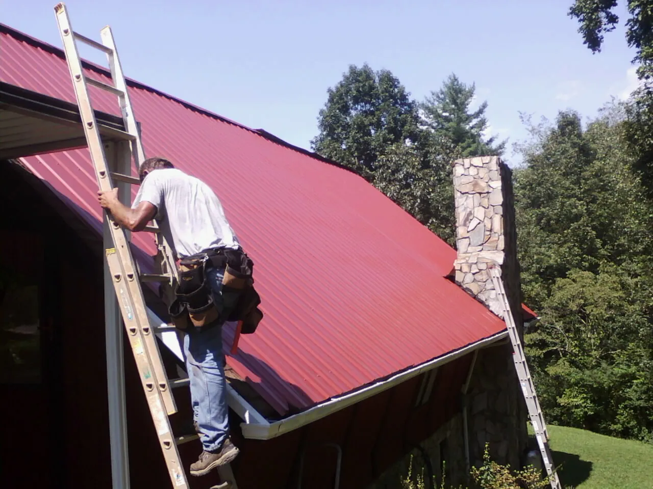 Roofer on a ladder beside a red metal roof and stone chimney — Soffit & Fascia Repair by Roofing Brothers in Bovill